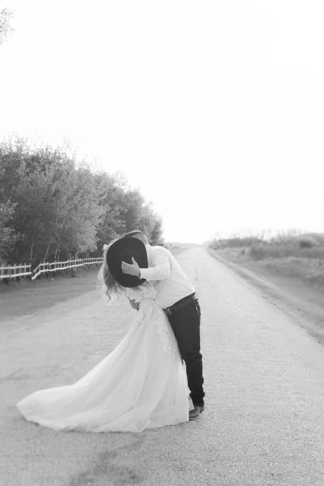 Bride and Groom share a dip kiss in the middle of a gravel road and are hidden behind a cowboy hat at Circle H Ranch in Saskatoon Saskatchewan. Photo Taken by Pulse Productions Inc.